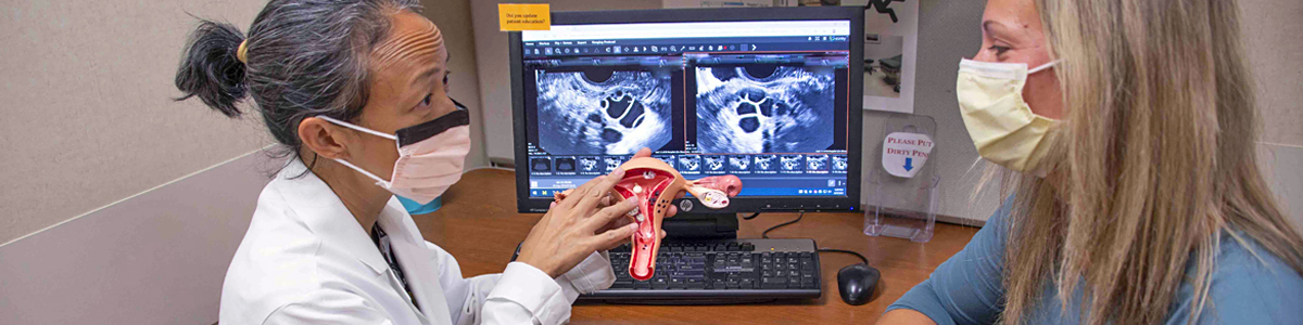 Asian female doctor wearing mask and white coat holding uterus model and speaking to female patient wearing mask, with computer monitor in background showing images